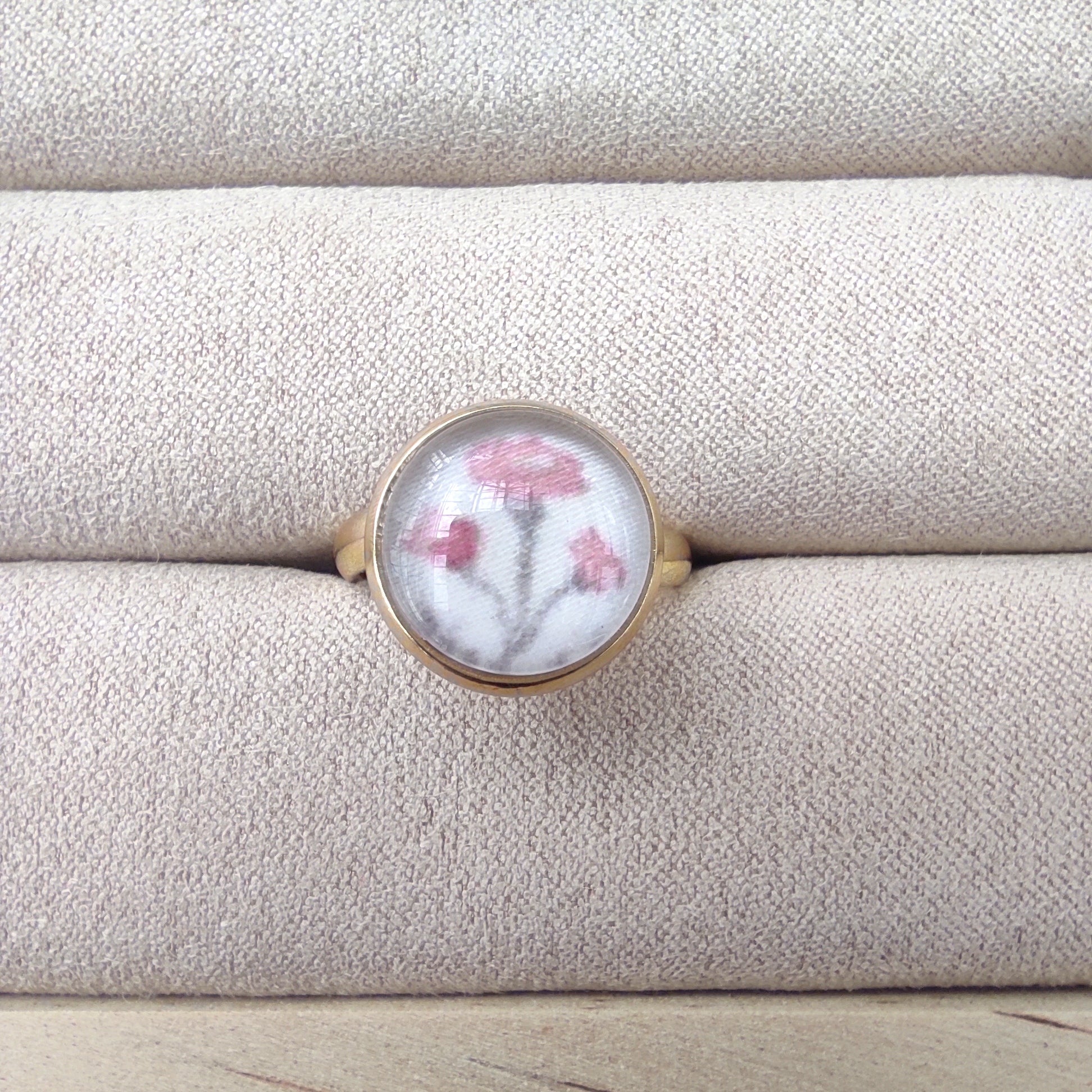Bague acier inoxydable réglable motif floral, motif feuillage. Une fleur rose est placée au centre de la bague qui est dorée. La bague est placée sur un présentoir.