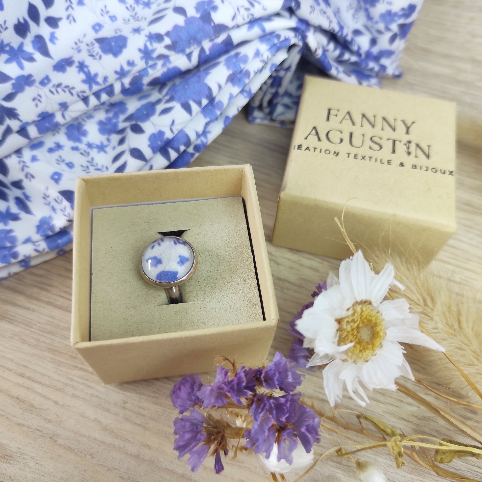 Bague acier inoxydable réglable motif fleurs bleues avec fond blanc. La bague est placée dans un coffret avec le tissu à côté qui a été incorporé sous la perle.