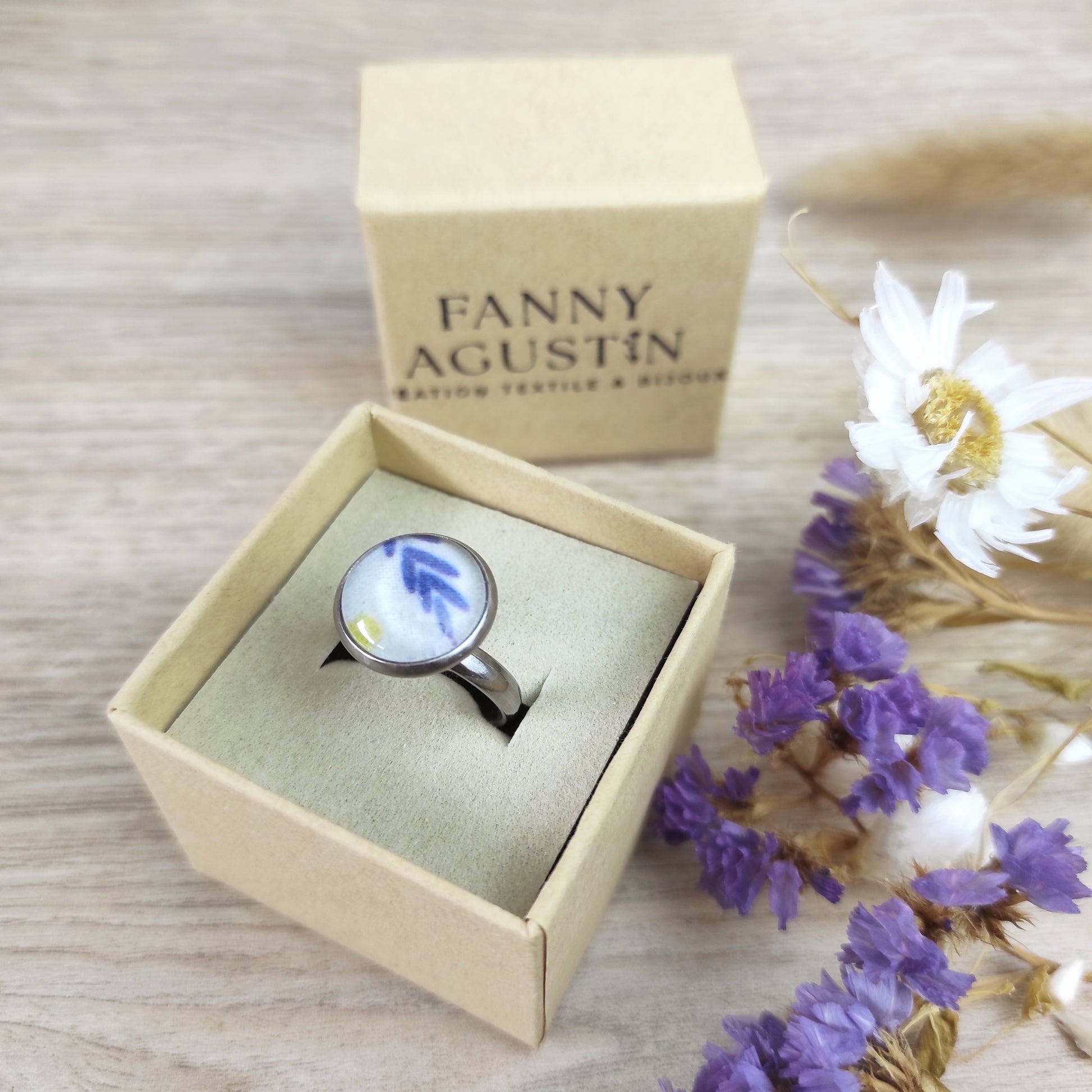 Bague acier inoxydable couleur argent réglable motif floral, motif fleurs mimosa. Une feuille stylisée bleue et un point jaune sont placés sur le tissu au centre de la perle.