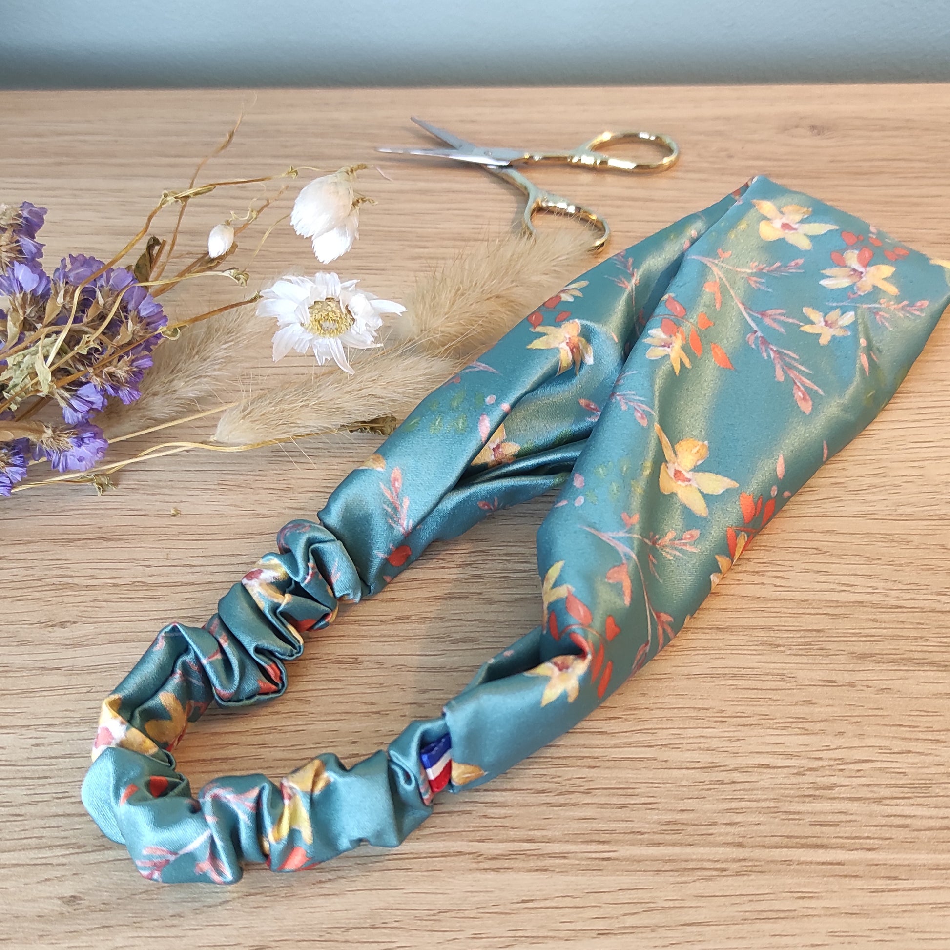 Accessoire pour cheveux, bandeau en satin avec motif fleuri dessiné par Fanny Agustin. Bandeau vert canard avec fleurs jaunes et feuillage marron et orange.