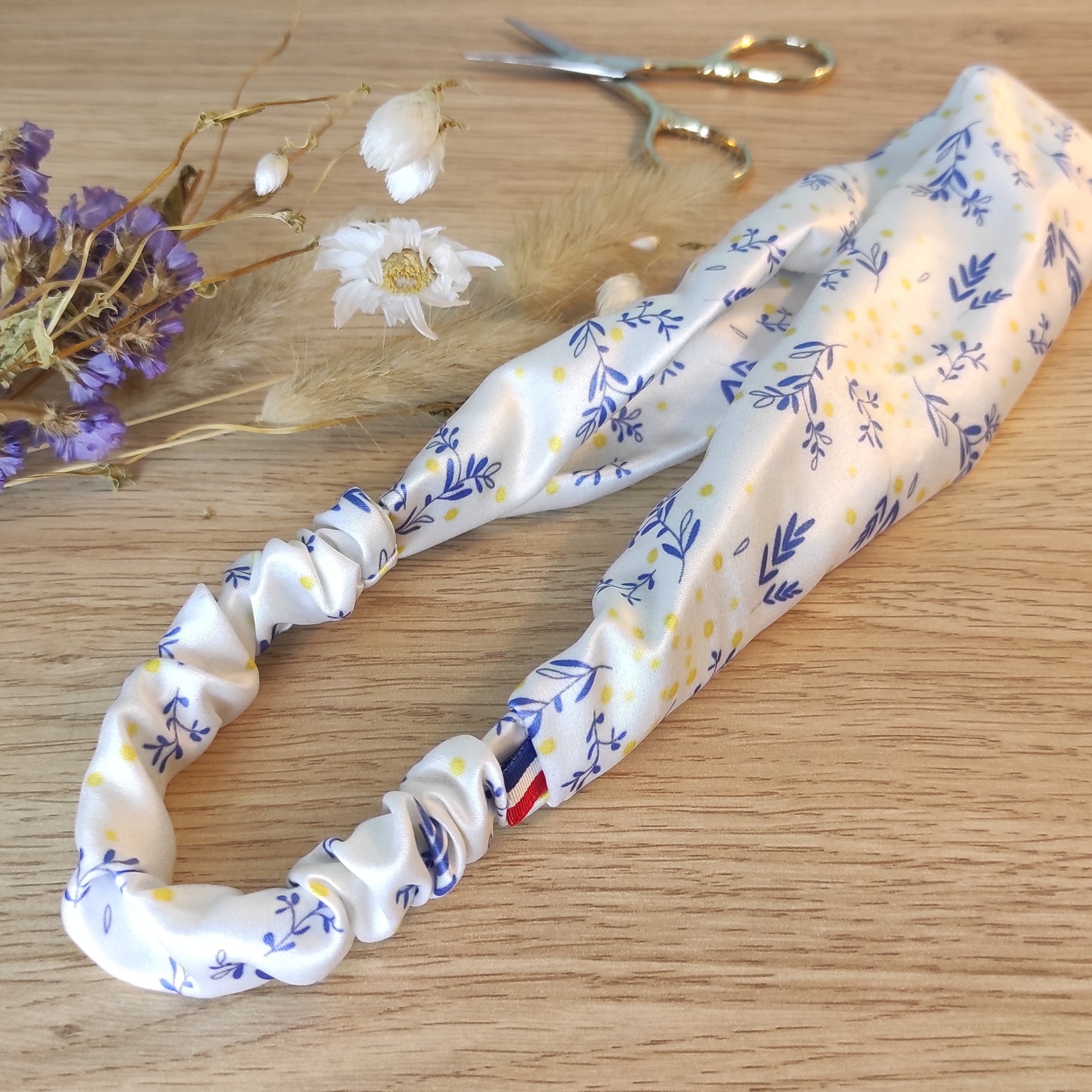 Accessoire pour cheveux, bandeau en satin avec motif fleuri dessiné par Fanny Agustin. Le bandeau pour cheveux est blanc, avec quelques touches de couleur jaune et bleu.