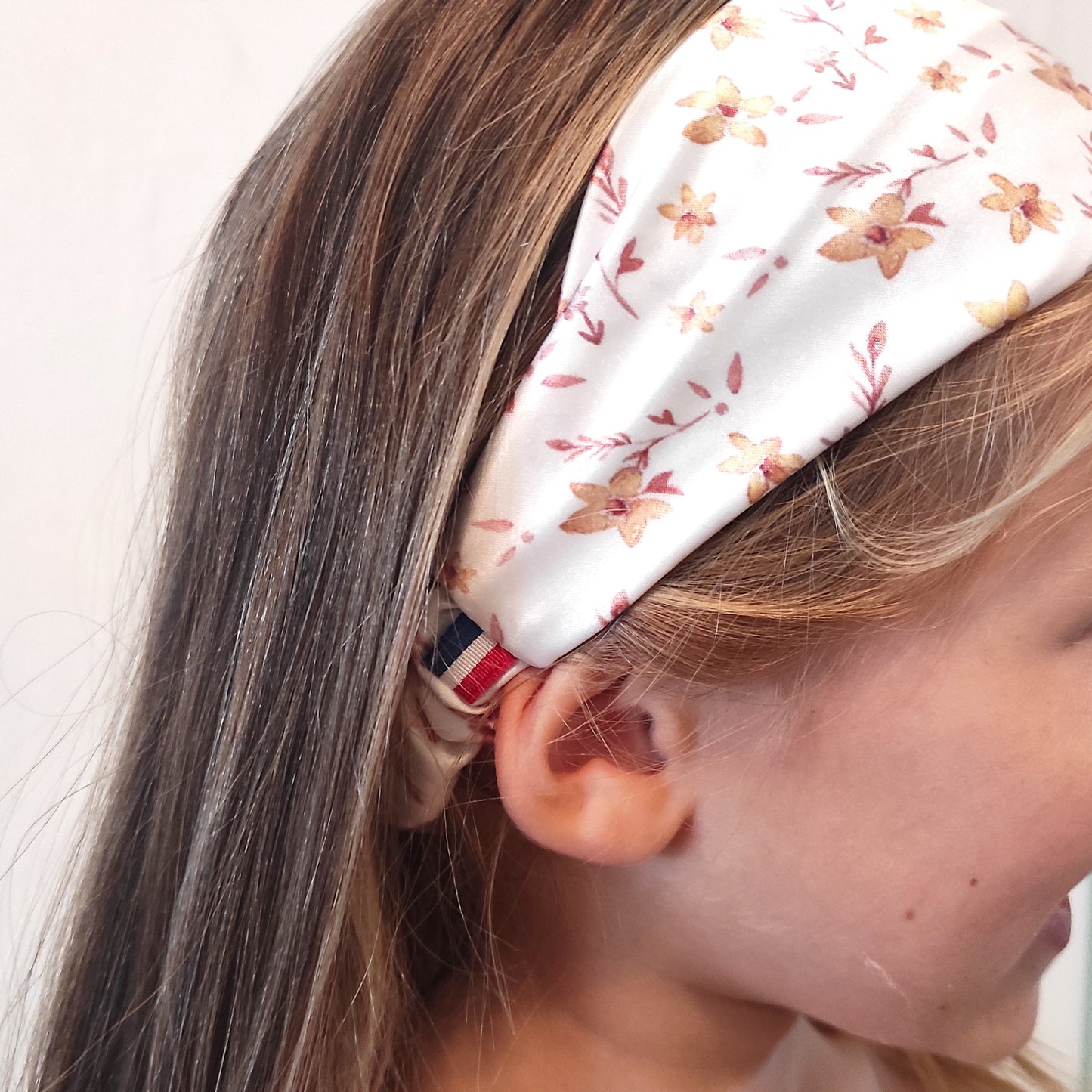 Bandeau au motif fleurs dorées porté sur fillette, le fond est blanc, et le motif est parsemé de fleurs jaunes et feuilles marrons. Un ruban bleu blanc rouge se trouve au dessus de l'oreille.