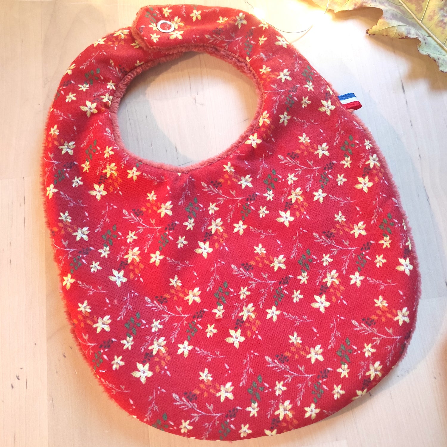 Bavoir au motif fleuri encre BIO, motif petites fleurs jaunes, feuilles marron, vertes et oranges, sur fond rouge.