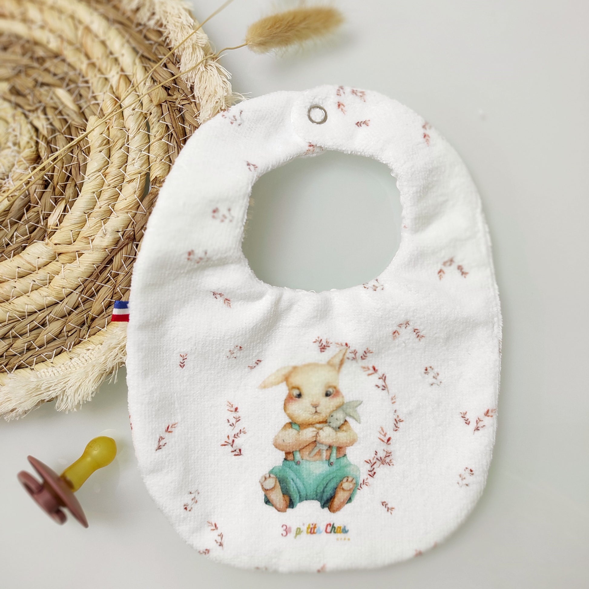 Bavoir avec motif lapin dessiné par Fanny Agustin. Le bavoir est blanc et le lapin porte une salopette turquoise, il est assis et sert un doudou lapin gris dans ses bras. Le bavoir est parsemé de petites feuilles couleur paprika. Le bavoir est réversible.