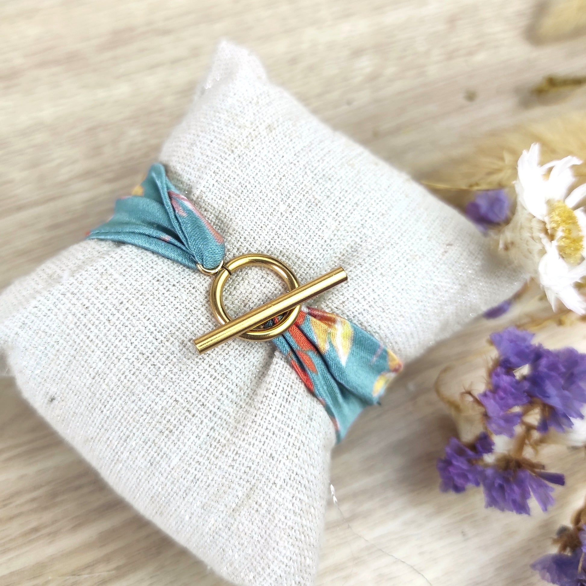 Bracelet en tissu avec motif fleuri dessiné par Fanny Agustin. Anneau fermoir en T couleur doré. Le tissu présente des fleurs jaunes et feuillage marron et orange.