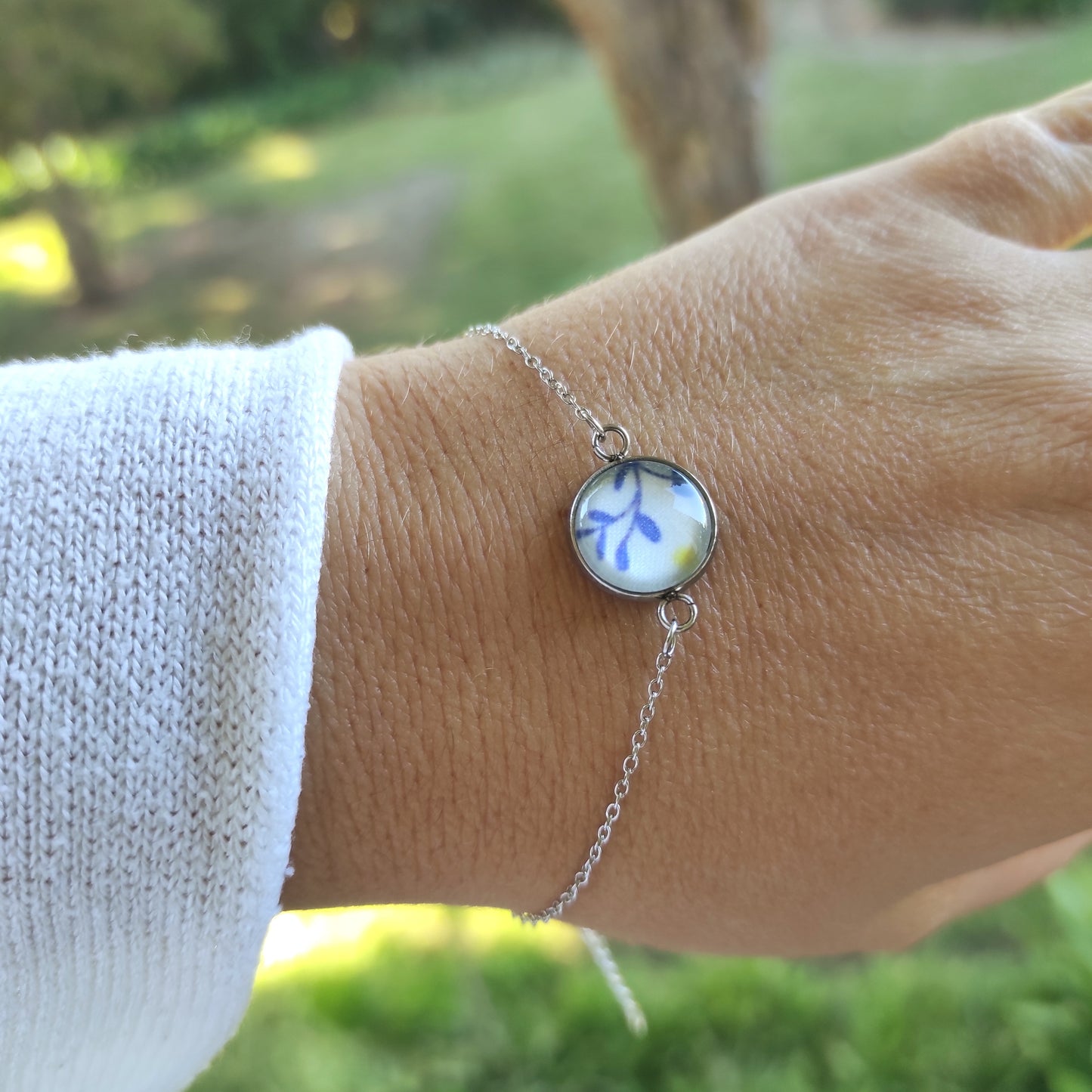 Bracelet réglable acier inoxydable couleur argent positionné sur un poignet. Le motif à l'intérieur comporte plusieurs un motif mimosa, feuilles bleues stylisées avec un point jaune rappelant le mimosa, avec un fond blanc. Perle de 12 mm de diamètre.
