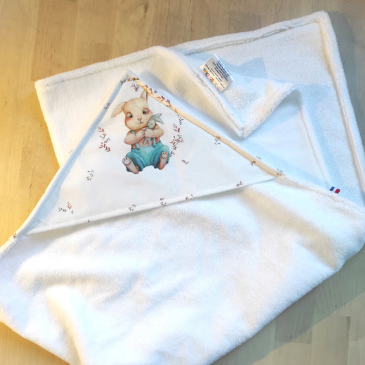 Cape de bain avec motif lapin sur la capuche. Le lapin porte un doudou lapin dans ses mains. La cape est blanche.