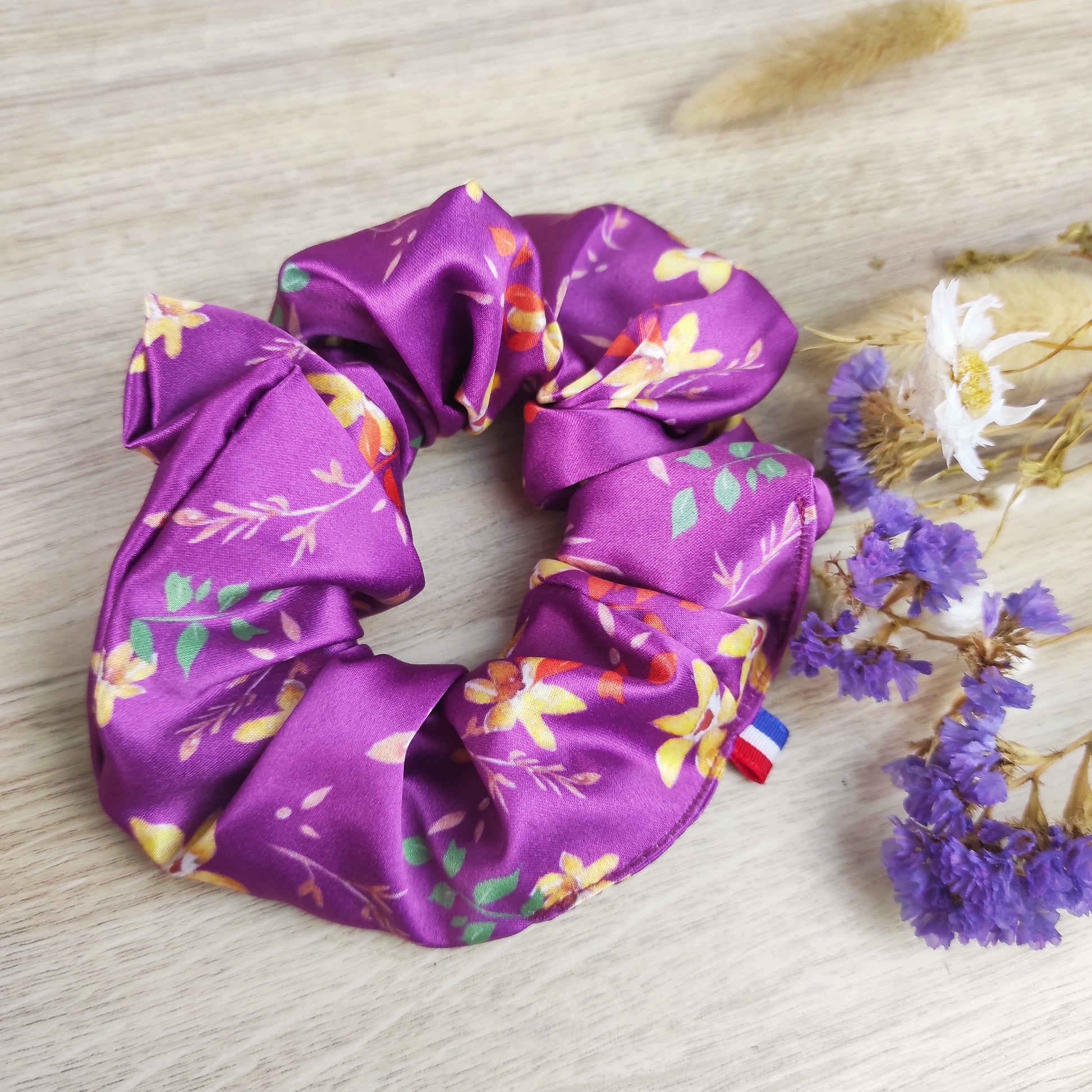 Accessoire cheveux en satin fleuri violet, motif dessiné par Fanny Agustin. Le chouchou comporte des fleurs jaunes, feuille oranges, marron et vertes, le fond est violet.