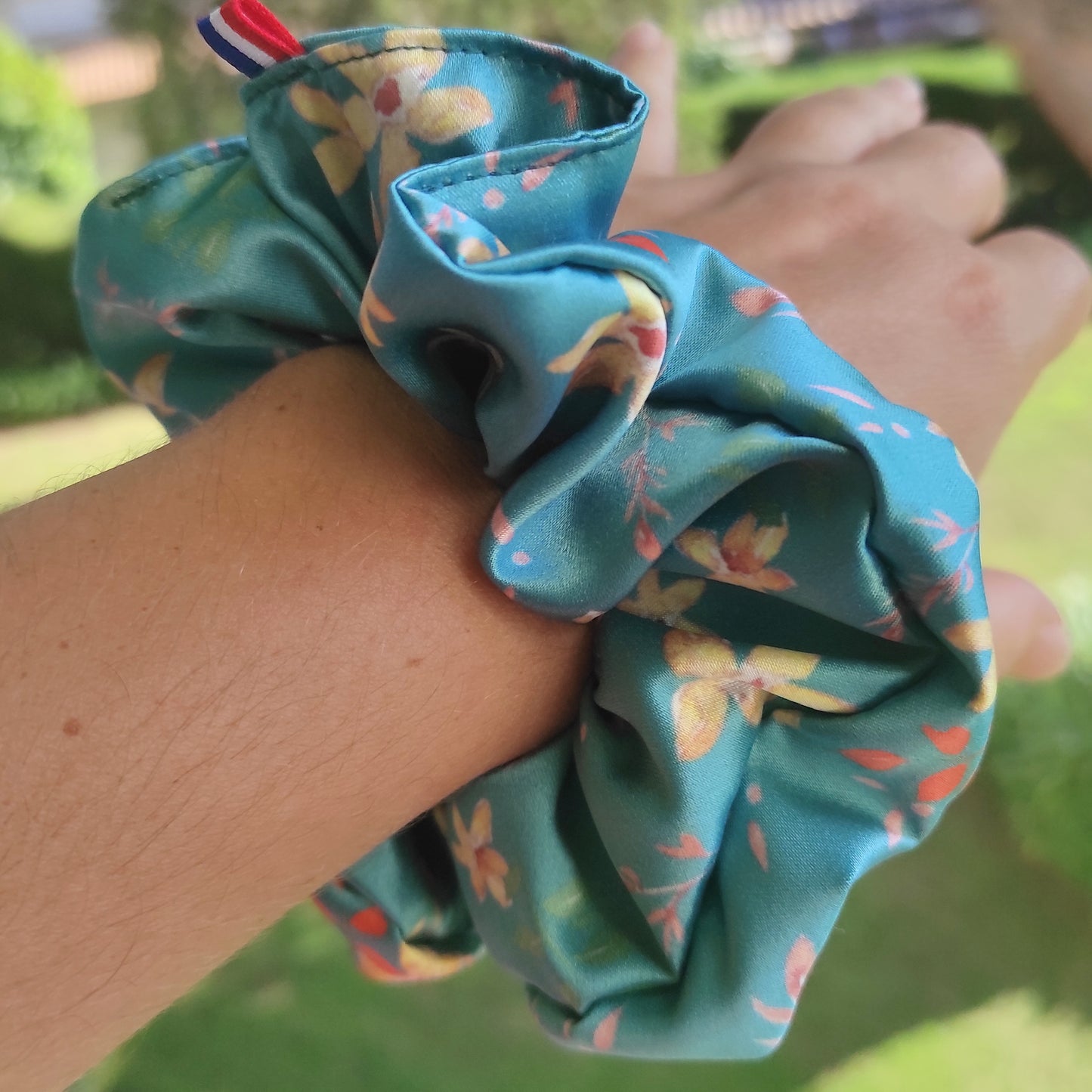 Gros chouchou porté au poignet en tissu satin fond vert canard et fleurs dorées, avec quelques touches de orange. Création Fanny Agustin, avec petit ruban Français bleu blanc rouge. 