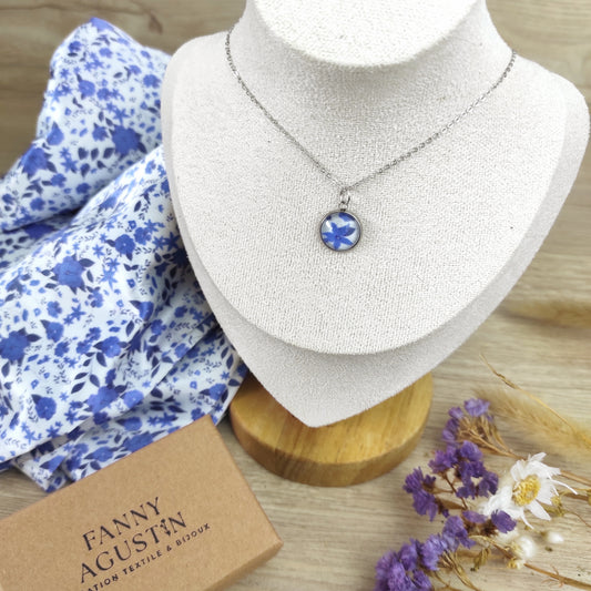 Collier acier inoxydable avec pendentif fleuri , motif fleurs bleues, avec fond blanc. Chaîne fine et pendentif de 12 mm, le collier est placé sur un présentoir avec un coffret à côté et le tissu dessiné par Fanny Agustin, incorporé à l'intérieur du bijou.