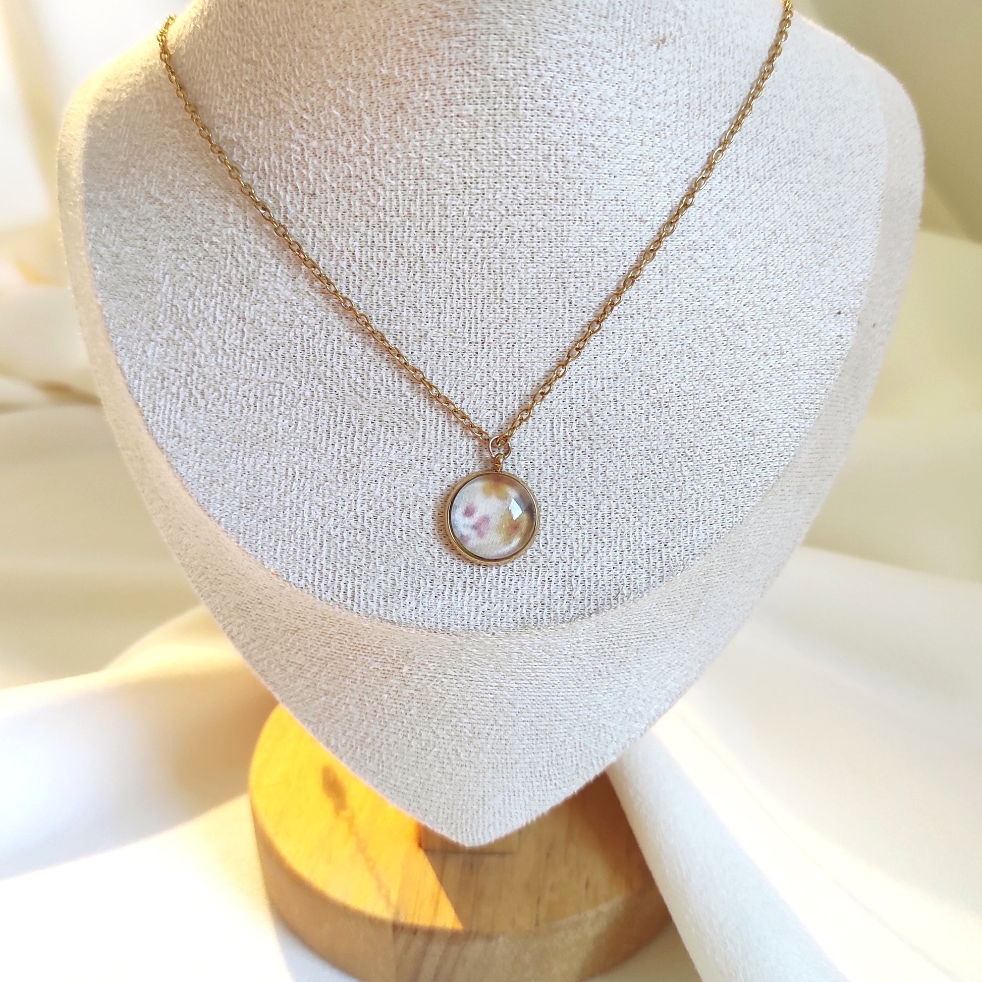 Collier doré avec perle de 12 mm et chaîne fine sur présentoir. Le motif est de petites fleurs dessinées en aquarelle et imprimé sur tissu, qui est ensuite incorporé dans la perle de verre. Il y a du jaune, rose, vert et fond blanc, motif délicat et discret. 