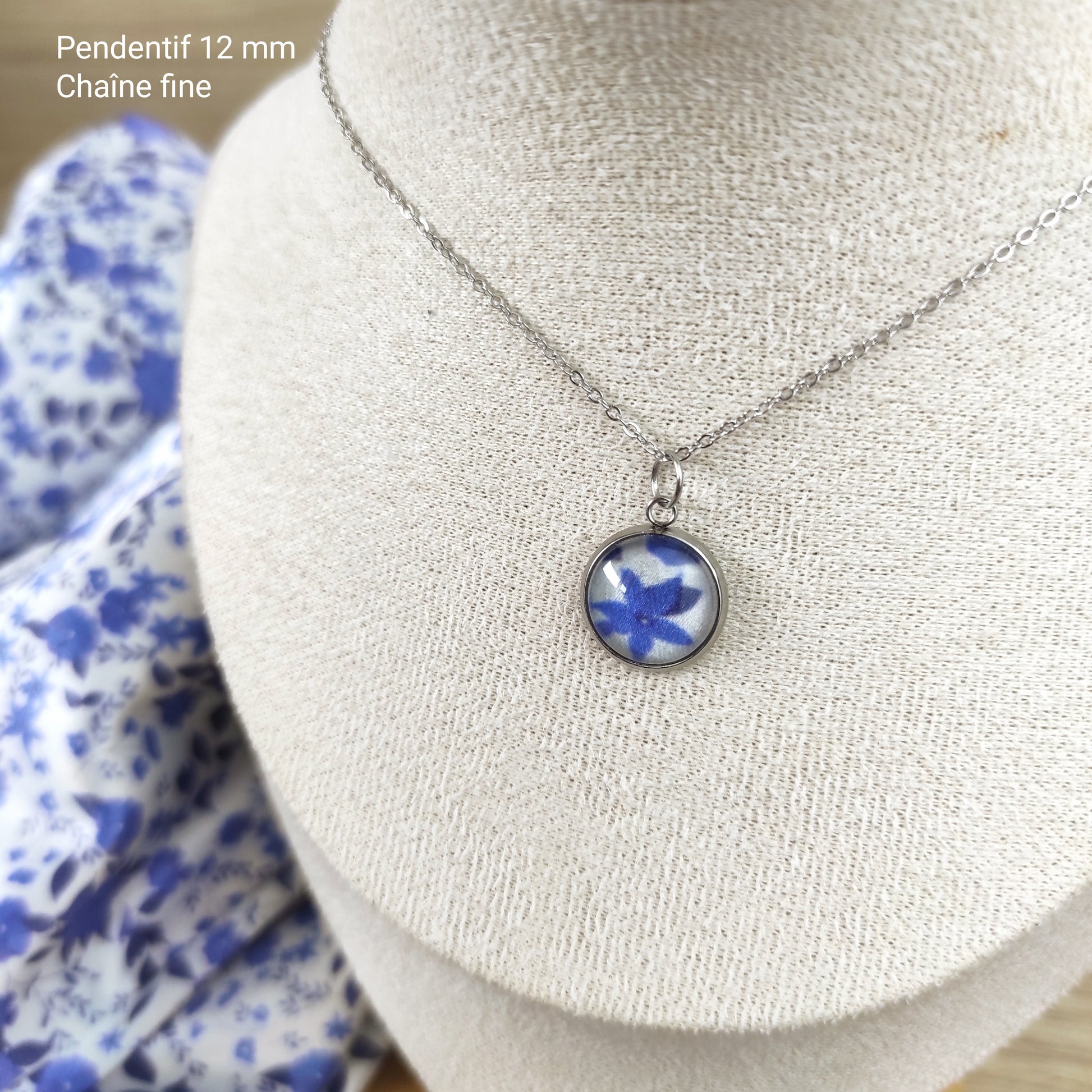 Collier acier inoxydable avec pendentif fleuri , motif fleurs bleues, avec fond blanc. Chaîne fine et pendentif de 12 mm, le collier est placé sur un présentoir  et le tissu dessiné par Fanny Agustin, incorporé à l'intérieur du bijou.