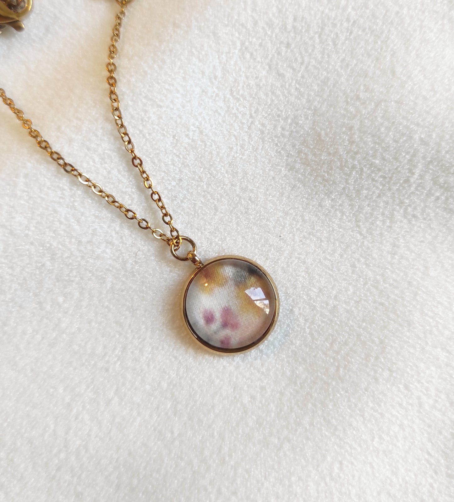 Collier doré avec perle de 12 mm et chaîne fine sur présentoir. Le motif est de petites fleurs dessinées en aquarelle et imprimé sur tissu. Il y a du jaune, rose, vert et fond blanc, motif délicat et discret. 