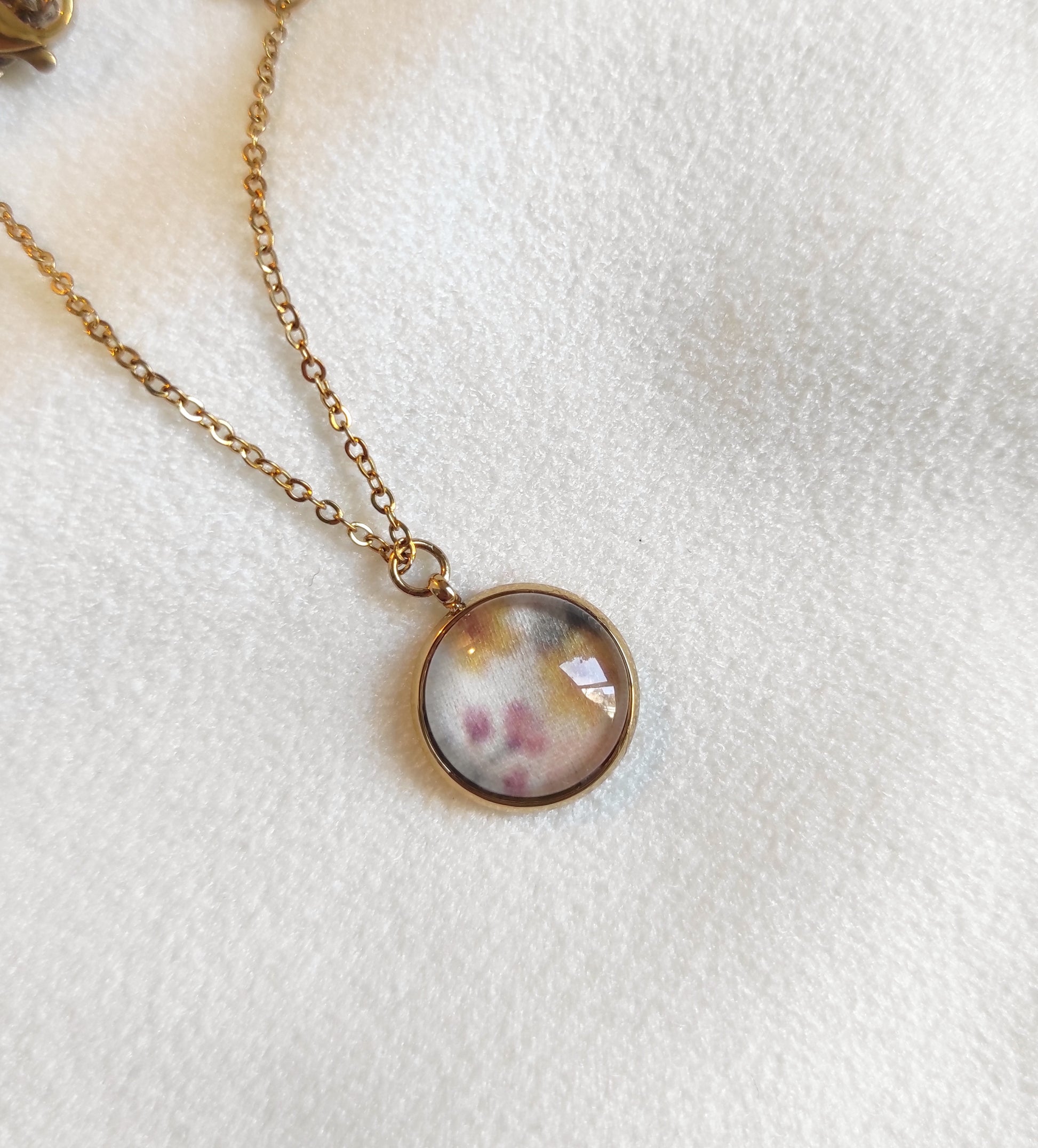 Collier doré avec perle de 12 mm et chaîne fine sur présentoir. Le motif est de petites fleurs dessinées en aquarelle et imprimé sur tissu. Il y a du jaune, rose, vert et fond blanc, motif délicat et discret. 