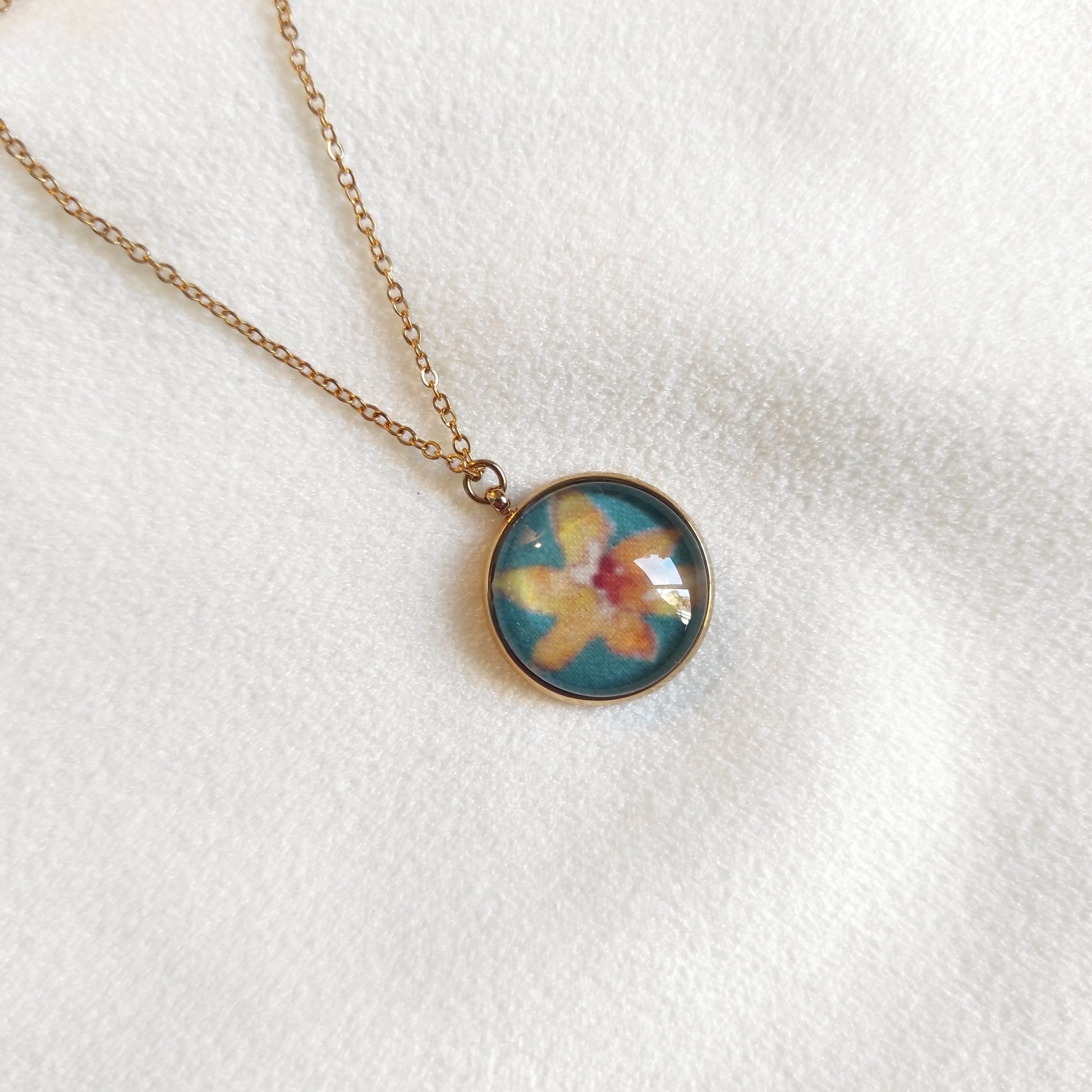 Collier doré avec perle de 12 mm et chaîne fine. Le motif est de petites fleurs dessinées et imprimé sur tissu. Il y a une fleur jaune au centre avec un fond vert. 