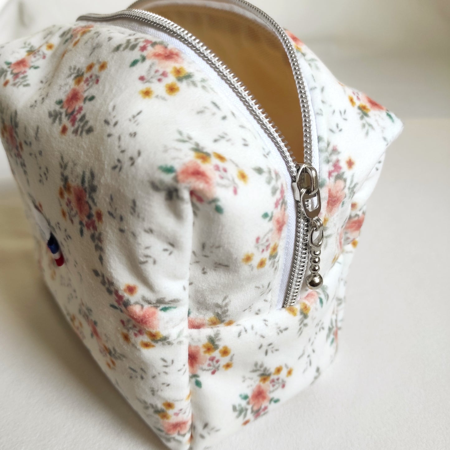 Trousse de toilette en velours doux imprimé localement et fabrication Française. Le dessin des fleurs est réalisé par Fanny Agustin ainsi que la fabrication à la main. 
La trousse en photographie est de taille normale, on peut y ajouter des accessoires à maquillage ou parfum comme représenté sur la photographie. Les motifs sont doux, avec du jaune, saumon et vert sur fond blanc. On voit la trousse de côté avec la fermeture éclair argentée.