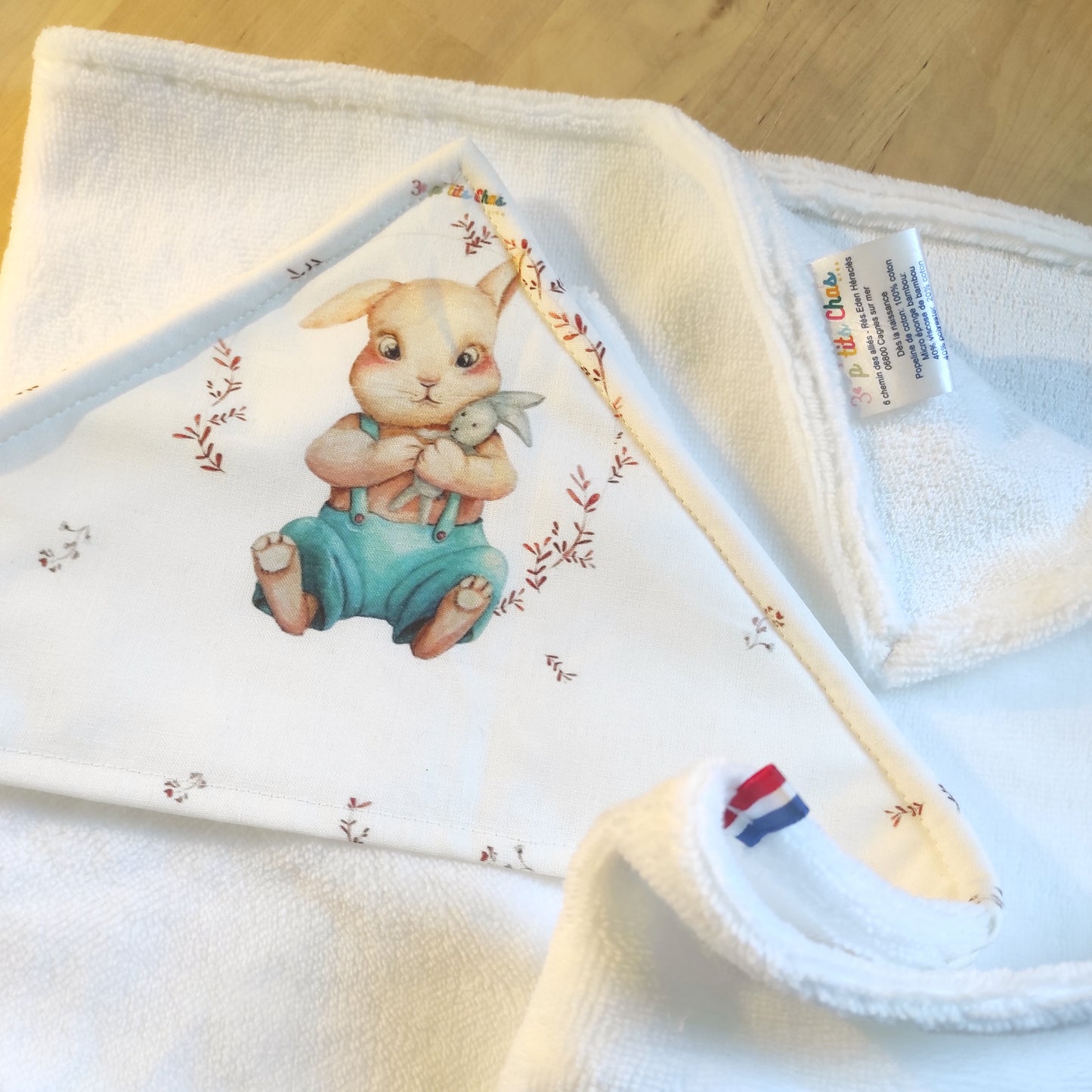 Détail capuche d'une cape de bain avec motif lapin sur la capuche. Le lapin porte un doudou lapin dans ses mains. La cape est blanche. Un ruban bleu blanc rouge est placé sur le côté.