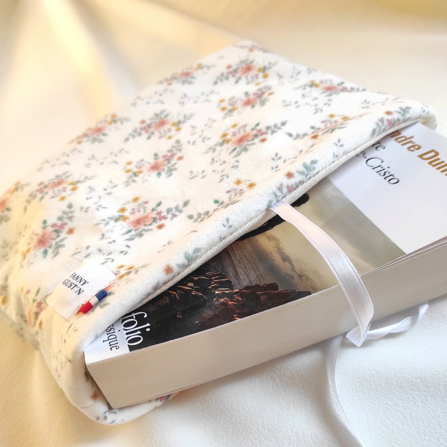 Pochette à livre en velours fleuri avec fleurs oranges, jaunes et roses et quelques feuilles vertes sur fond blanc. Le motif est dessiné par Fanny Agustin, puis fabriqué artisanalement à Cagnes-sur-Mer en France. Sur la photo, on voit que la pochette contient un livre de poche assez volumineux.
