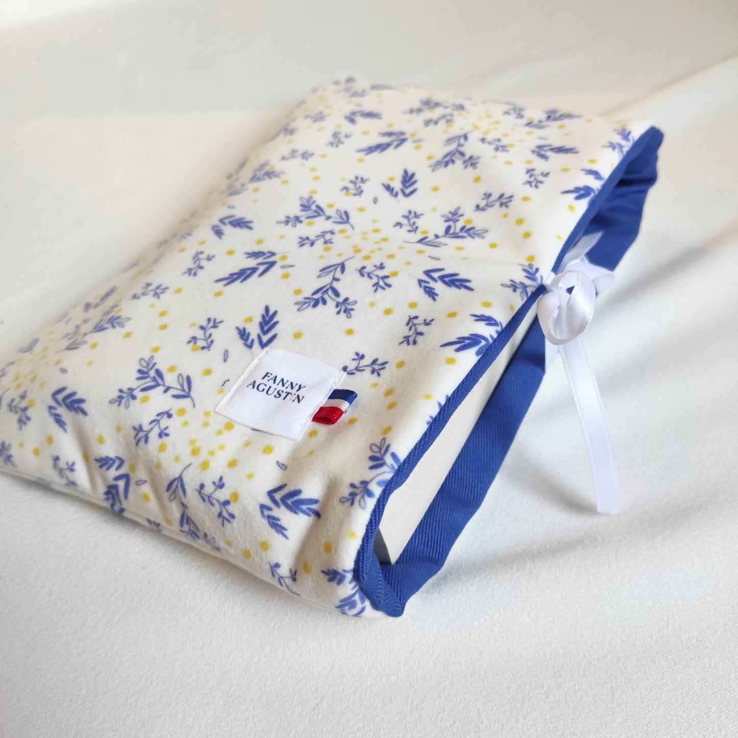 Pochette à livre, format poche, en velours fleuri, imprimé localement, fabrication Française et artisanale. Le motif est dessiné par Fanny Agustin et la pochette est fabriquée par ses soins à la main. Le motif sur la photographie est mimosa, feuilles stylisées bleues avec quelques points jaunes sur fond blanc. La doublure est bleue et rappelle la couleur des motifs. La pochette se referme avec un ruban en satin blanc.