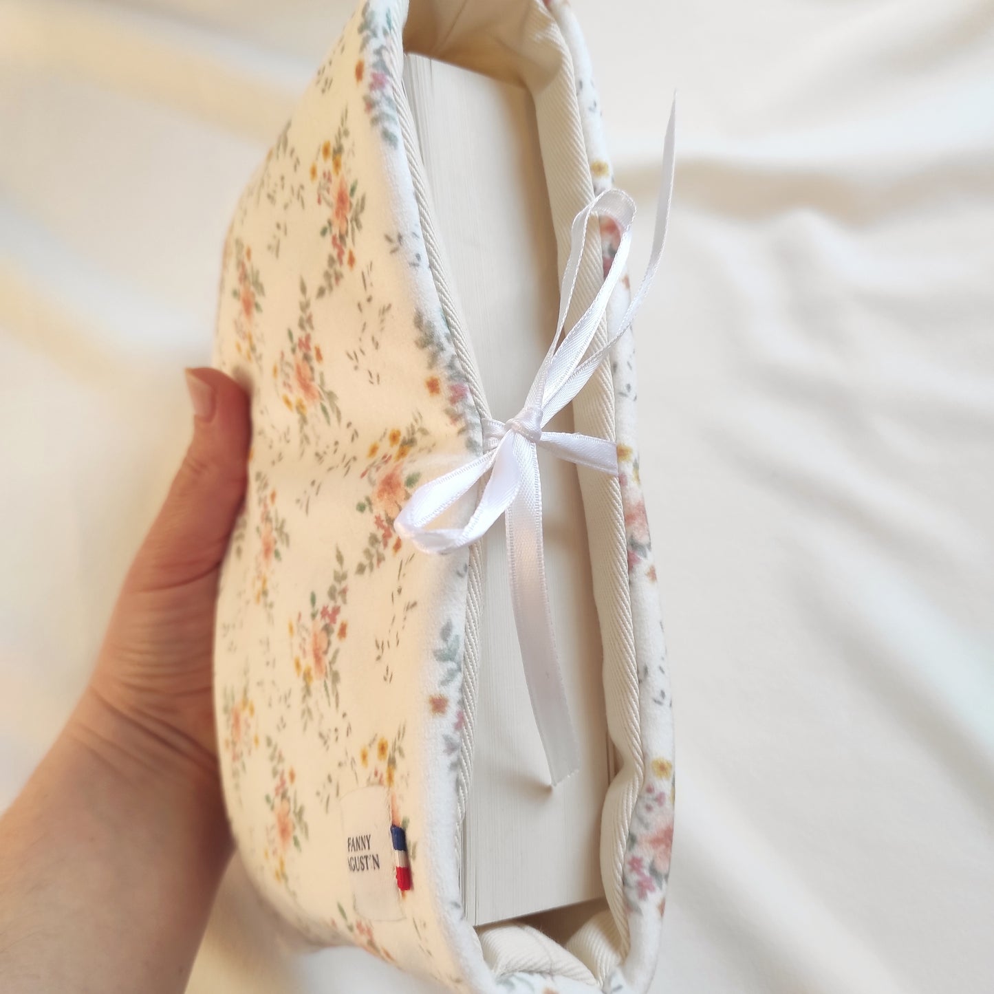 Pochette à livre en velours fleuri avec fleurs oranges, jaunes et roses et quelques feuilles vertes sur fond blanc. Le motif est dessiné par Fanny Agustin, puis fabriqué artisanalement à Cagnes-sur-Mer en France. Sur la photo, la pochette est fermée avec un ruban en satin blanc.