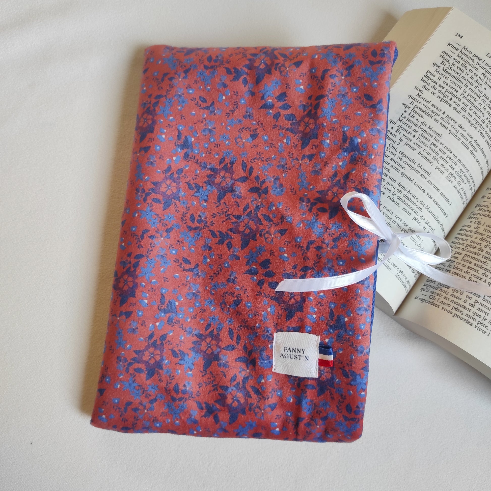 Pochette à livre, format poche, en velours fleuri, imprimé localement, fabrication Française et artisanale. Le motif est dessiné par Fanny Agustin et la pochette est fabriquée par ses soins à la main. Le motif sur la photographie est fleurs bleues sur fond paprika. La pochette se referme avec un ruban en satin blanc.