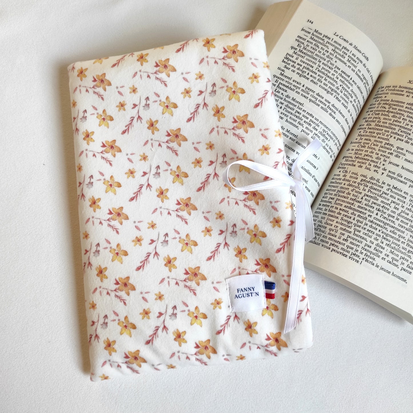 Pochette à livre, format poche, en velours fleuri, imprimé localement, fabrication Française et artisanale. Le motif est dessiné par Fanny Agustin et la pochette est fabriquée par ses soins à la main. Le motif sur la photographie est fleurs dorées, les fleurs sont jaunes et blanches avec quelques feuilles marrons sur fond blanc. La pochette se referme avec un ruban en satin blanc.
