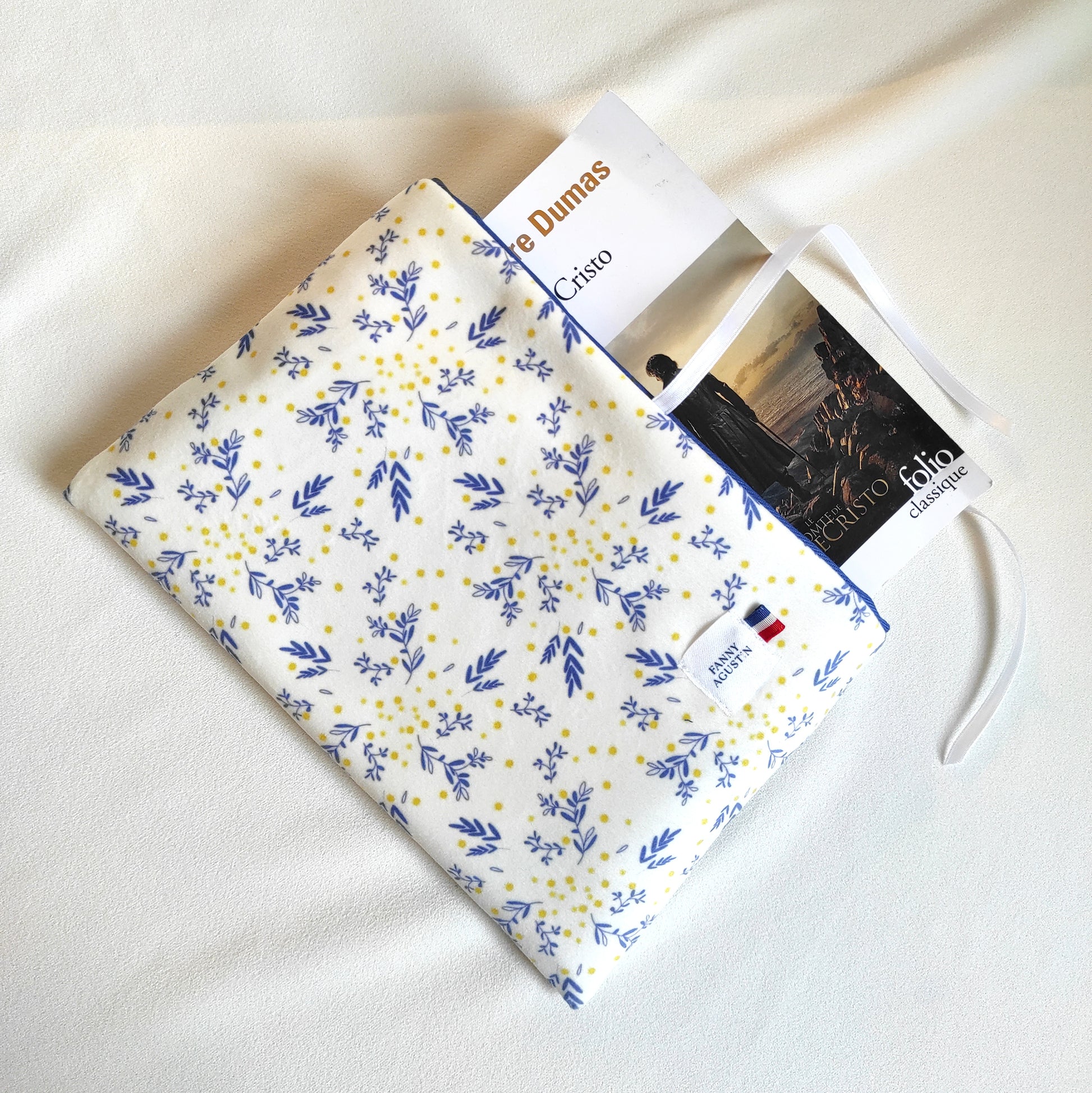 Pochette à livre, format poche, en velours fleuri, imprimé localement, fabrication Française et artisanale. Le motif est dessiné par Fanny Agustin et la pochette est fabriquée par ses soins à la main. Le motif sur la photographie est mimosa, feuilles stylisées bleues avec quelques points jaunes sur fond blanc. La doublure est bleue et rappelle la couleur des motifs. La pochette se referme avec un ruban en satin blanc.
Un livre gros format est placé dans la pochette.