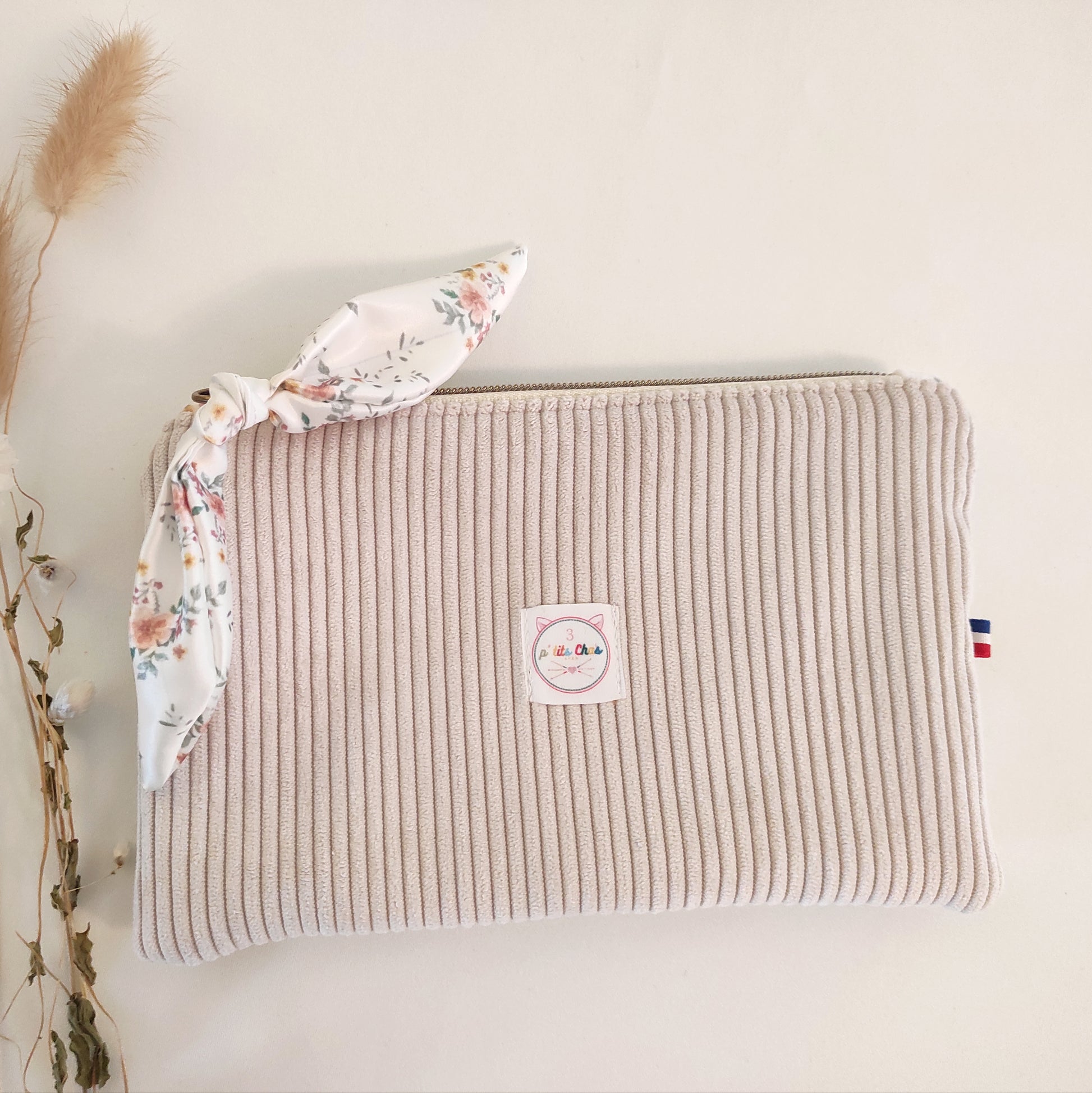 Pochette en velours écru, avec ruban fleuri et logo placé au centre. Détail du ruban made in France sur le côté.