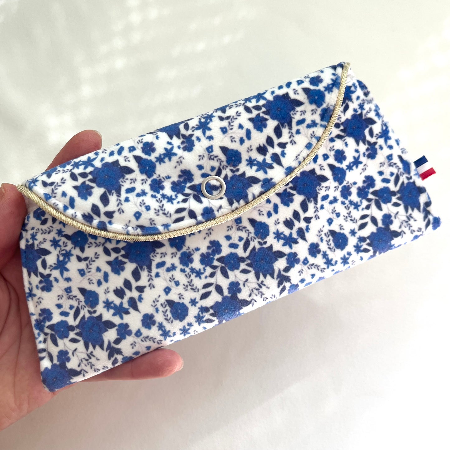 Pochette à lunettes en velours imprimé, motif fleurs bleues avec fond blanc dessiné par Fanny Agustin et fabrication artisanale Française. Un ruban doré apporte une finition plus chic.