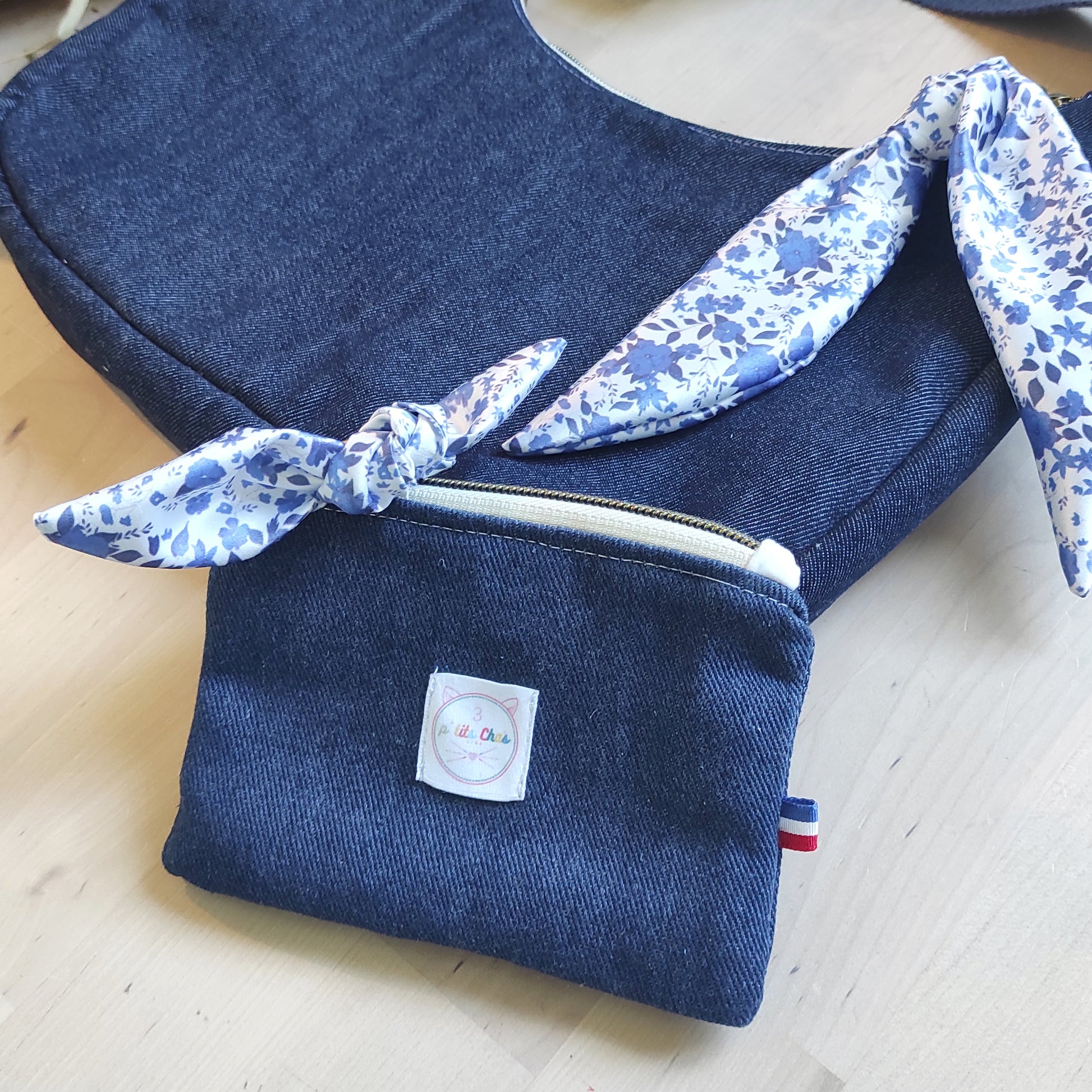 Porte monnaie en jean bleu foncé, avec son nœud en satin, placé devant un sac à main dans la même collection.