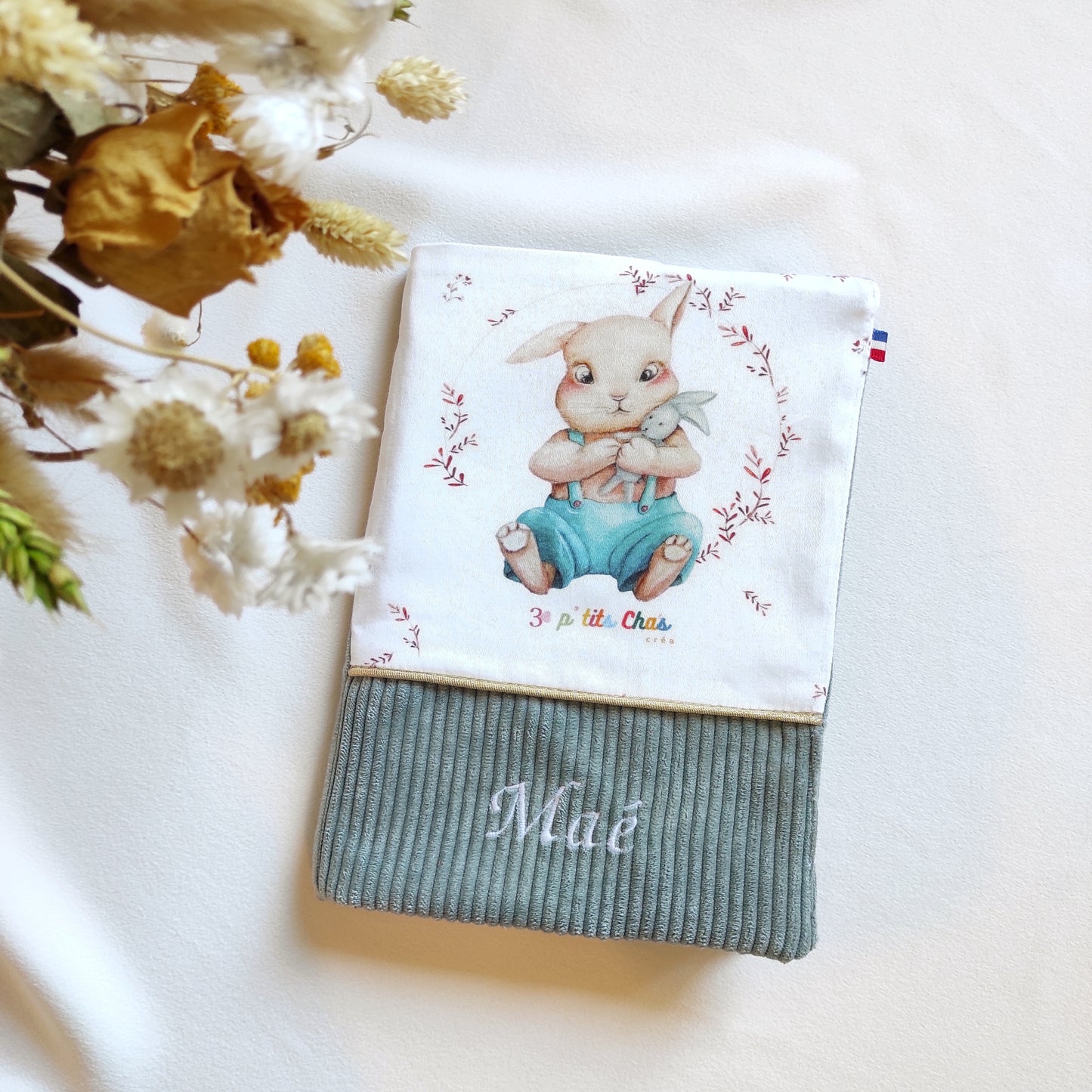 Protège carnet de santé personnalisé. Sur la photo, on trouve un carnet avec motif lapin et velours vert de gris, ruban doré et prénom brodé en blanc sur le velours. 