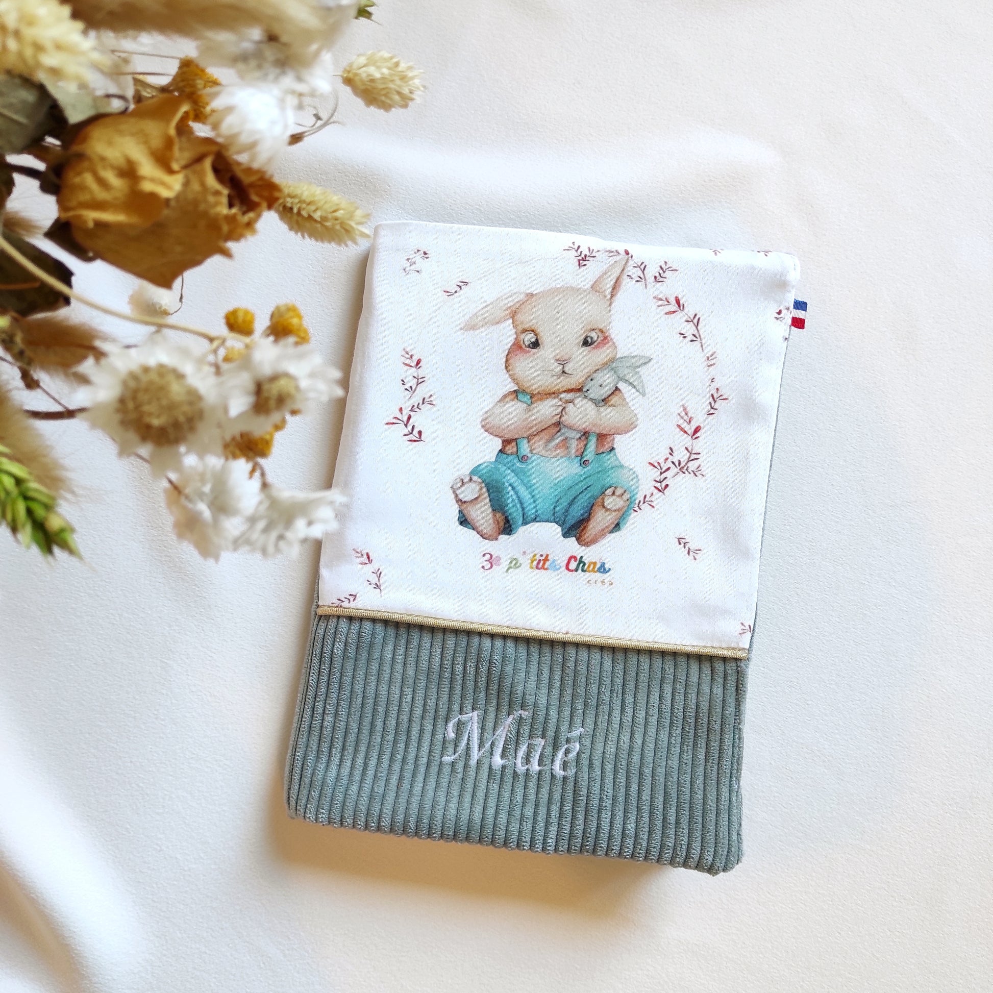 Protège carnet de santé personnalisé. Sur la photo, on trouve un carnet avec motif lapin et velours vert de gris, ruban doré et prénom brodé en blanc sur le velours. 