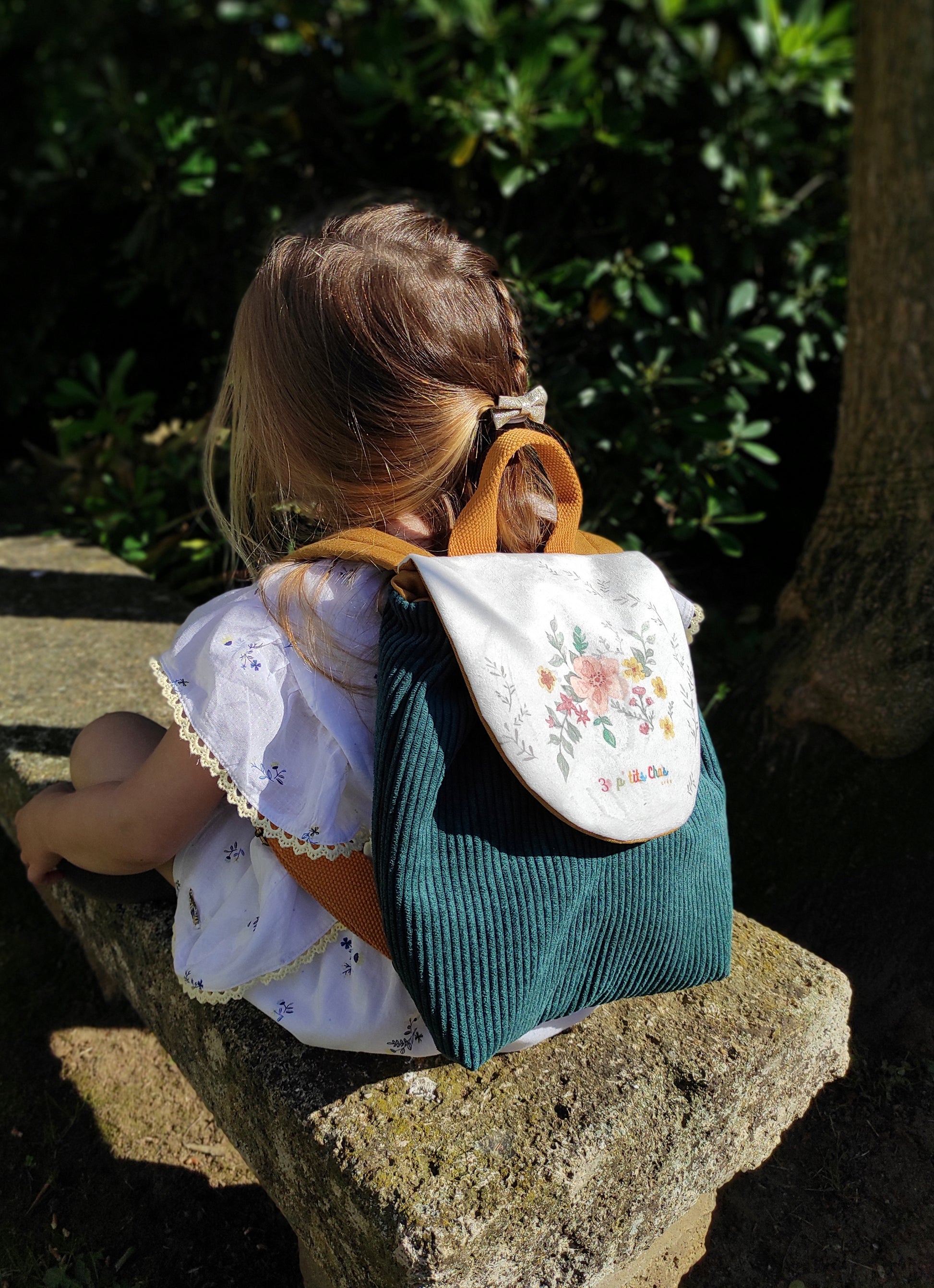 Motif grosses fleurs aquarelle sur fond blanc, avec couleur du sac vert canard. Une fillette assise sur un banc porte le sac sur son dos.