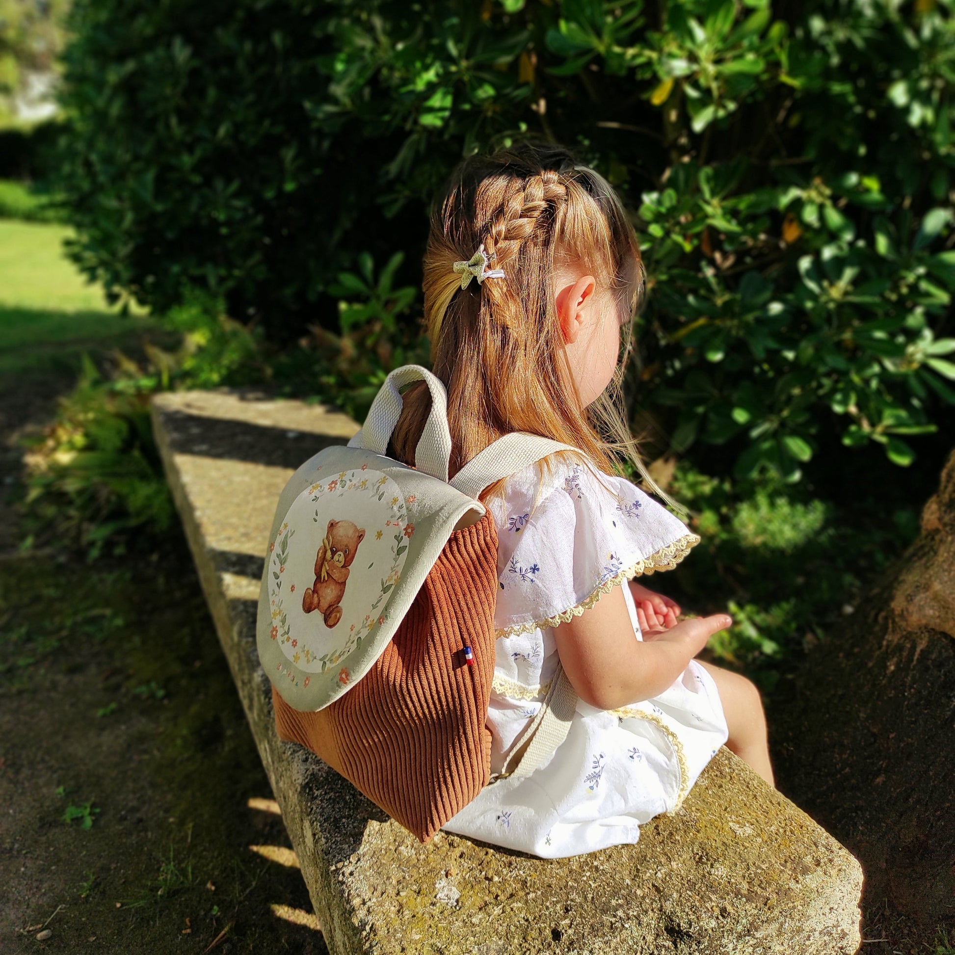 Sac à dos ourson vert avec velours marron, porté sur fillette assise sur un banc.