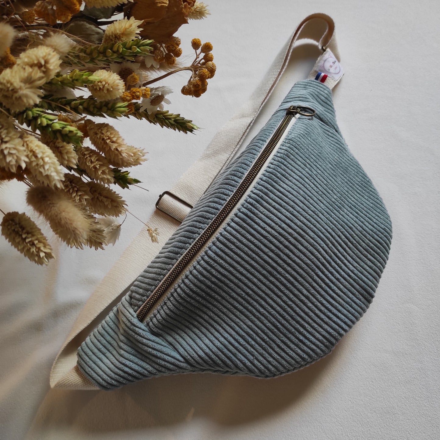 Banane placée sur une table, couleur vert de gris, avec fermeture éclaire écrue et bronze. Détail sur le côté du logo et ruban made in France.