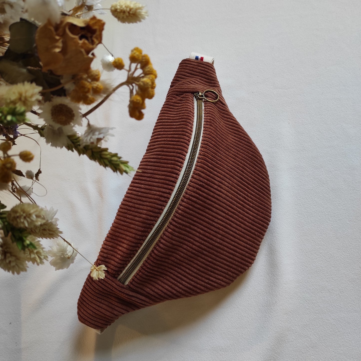 Banane placée sur une table couleur marron, avec fermeture éclaire écrue et bronze. Détail sur le côté du logo et ruban made in France.