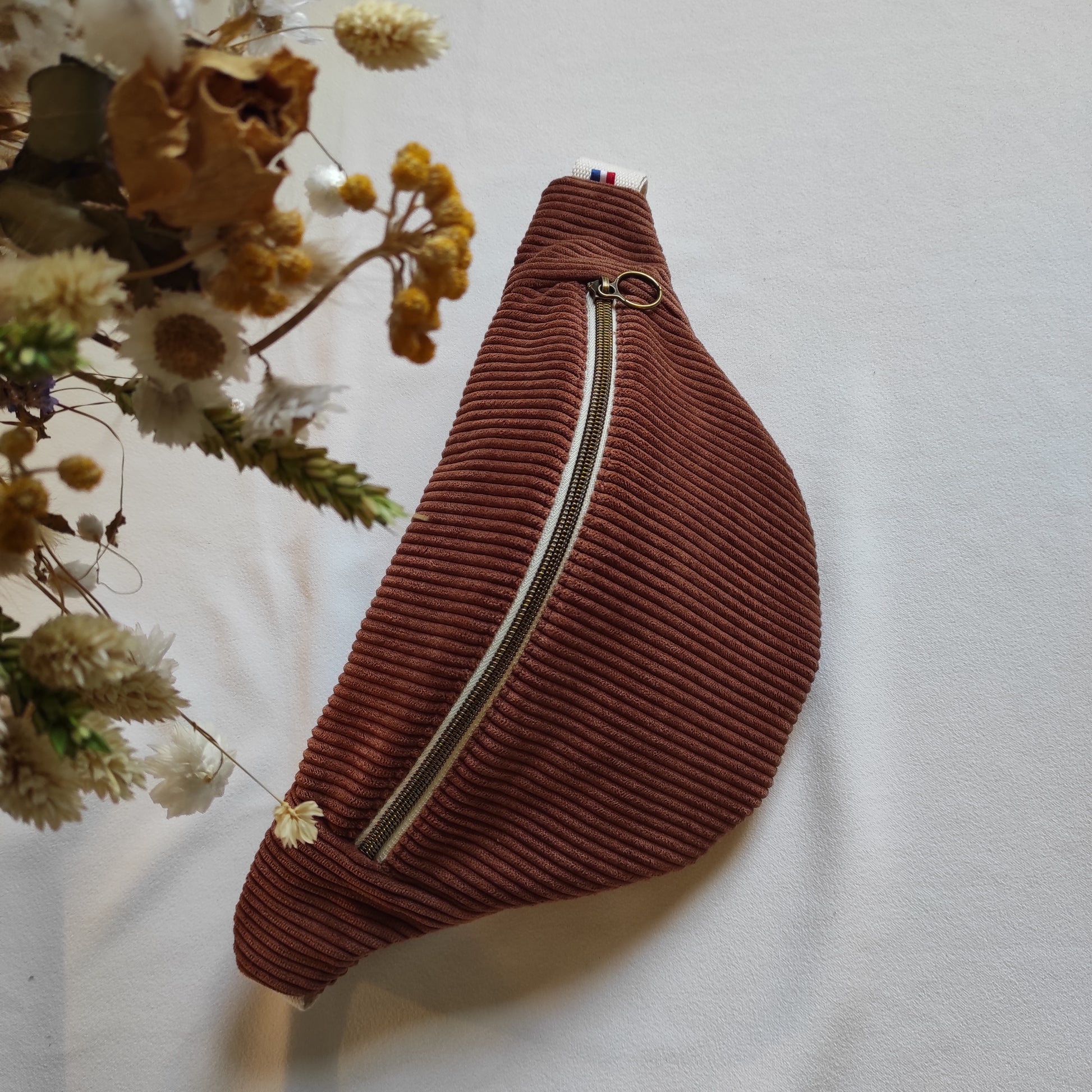 Banane placée sur une table couleur marron, avec fermeture éclaire écrue et bronze. Détail sur le côté du logo et ruban made in France.